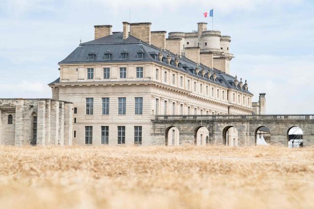 This photograph shows dried out cut grass with the Vincennes Castle and a fluttering French flag in the background at the Vincennes Woods, a park in eastern Paris, on July 13, 2026, amid a heatwave. The Paris region, and large parts of the rest of France, is currently experiencing the third heatwave since May 2026, increasing the risk of fires. The three heatwaves have seen temperature records broken in several countries across Europe and have caused thousands of excess deaths, according to estimates in Belgium, Britain, France and Spain. (Photo by Martin LELIEVRE / AFP)