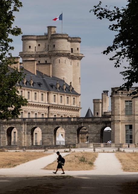 A photographer walks past the Vincennes Castle with a fluttering French flag and next to dried out cut grass at the Vincennes Woods, a park in eastern Paris, on July 13, 2026, amid a heatwave. The Paris region, and large parts of the rest of France, is currently experiencing the third heatwave since May 2026, increasing the risk of fires. The three heatwaves have seen temperature records broken in several countries across Europe and have caused thousands of excess deaths, according to estimates in Belgium, Britain, France and Spain. (Photo by Martin LELIEVRE / AFP)
