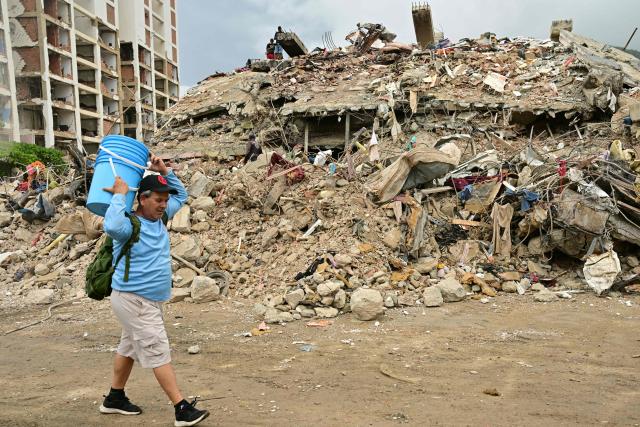 A man carries buckets past the rubble of a collapsed building in Caraballeda, La Guaira state, Venezuela, on July 13, 2026, following twin earthquakes that struck the region. The official death toll from Venezuela's devastating twin earthquakes rose to nearly 4,500 on July 12, 2026, as the government reported more than 19,500 people were now living in camps due to the destruction. (Photo by MARTIN BERNETTI / AFP)