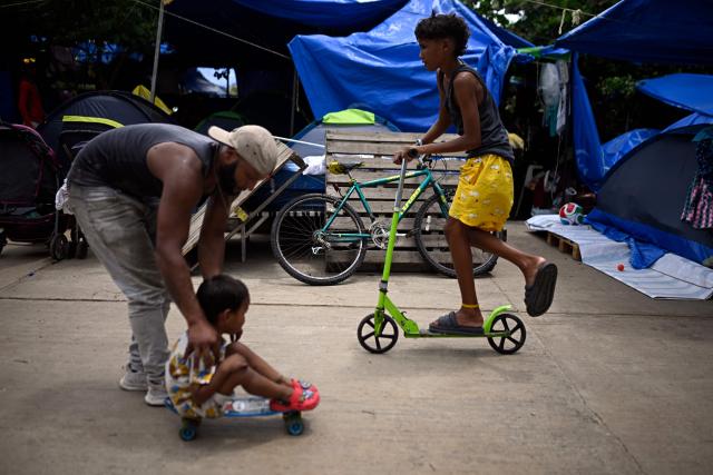 A boy rides a scooter as an adult helps a child on a skateboard at a makeshift camp housing people displaced by the twin earthquakes in Maiquetia, La Guaira state, Venezuela, on July 13, 2026. The death toll from the twin earthquakes that struck Venezuela nearly three weeks ago has surpassed 4,500, while the number of injured remains at about 17,000, according to the latest government report released on July 13. (Photo by Federico PARRA / AFP)