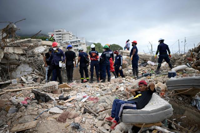 Rescue workers search through the rubble of a collapsed building as a resident sits on a mattress in Los Corales, La Guaira state, Venezuela, on July 13, 2026, following twin earthquakes that struck the region. The death toll from the twin earthquakes that struck Venezuela nearly three weeks ago has surpassed 4,500, while the number of injured remains at about 17,000, according to the latest government report released on July 13. (Photo by Federico PARRA / AFP)