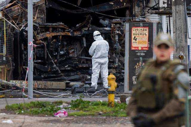 A police officer stands guard as a forensic expert inspects a store caght by fire after a van collided with a Metropolitan Transport Network (RED) bus in the Franklin neighborhood, Santiago, Chile, on July 14, 2026. Five people died in an accident between a van and a bus which caused a fire that engulfed the public transport vehicle and commercial premises. (Photo by Javier TORRES / AFP)