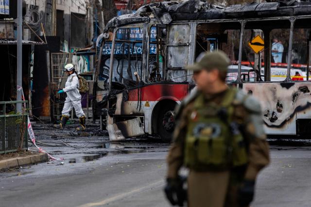 A policeman stands guard as a forensic expert walks next to a Metropolitan Transport Network (RED) bus after it collided with a van in the Franklin neighborhood, Santiago, Chile, on July 14, 2026. Five people died in an accident between a van and a bus which caused a fire that engulfed the public transport vehicle and commercial premises. (Photo by Javier TORRES / AFP)