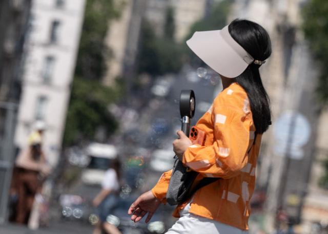 A pedestrian walks with an electronic handfan amid a heatwave in Paris on July 14, 2026. The Paris region, and large parts of the rest of France, is currently experiencing the third heatwave since May 2026, increasing the risk of fires. The three heatwaves have seen temperature records broken in several countries across Europe and have caused thousands of excess deaths, according to estimates in Belgium, Britain, France and Spain. (Photo by Martin LELIEVRE / AFP)