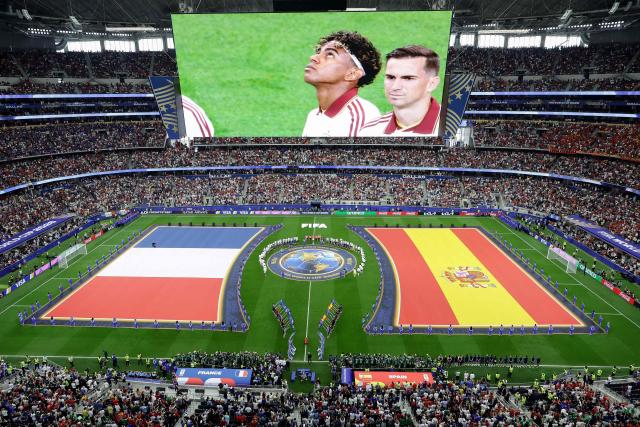 A jumbotron shows Spain's forward #19 Lamine Yamal as Frances' players (L) and Spain's players (R) stand for the national anthems ahead of the 2026 World Cup football tournament semi-final match between France and Spain at the Dallas Stadium in Arlington on July 14, 2026.  (Photo by Aric Becker / AFP)