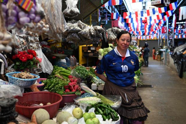 Teresa Hernandez, 50, poses for a picture wearing France's football team jersey at her vegetables stall in the Duenas Market where vendors show their support for their favourite team in the FIFA World Cup semifinals between France and Spain, in Santa Tecla, El Salvador, on July 14, 2026. As the tournament reaches its final stages, many market vendors have openly embraced their favorite teams, decorating stalls and market aisles with flags, jerseys and colors in celebration of the competing nations. (Photo by Marvin RECINOS / AFP)