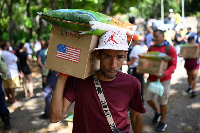 A resident carries an aid box bearing a US flag and a sack of rice on his shoulder as residents receive aid distributed by the Global Environment Mission (GEM) in Caraballeda, La Guaira state, Venezuela, on July 14, 2026, following twin earthquakes that struck the region. The death toll from the earthquakes that struck Venezuela almost three weeks ago has surpassed 4,700, while almost 17,000 people have been injured, an official report released on July 14 stated. (Photo by Federico PARRA / AFP)