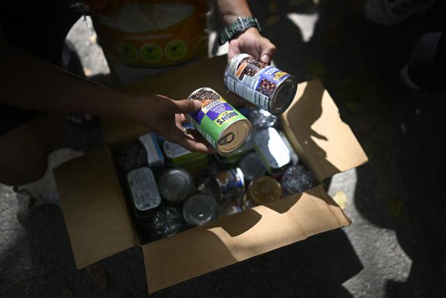 A person holds canned food over a box containing assorted supplies as residents receive aid distributed by the Global Environment Mission (GEM) in Caraballeda, La Guaira state, Venezuela, on July 14, 2026, following twin earthquakes that struck the region. The death toll from the earthquakes that struck Venezuela almost three weeks ago has surpassed 4,700, while almost 17,000 people have been injured, an official report released on July 14 stated. (Photo by Federico PARRA / AFP)