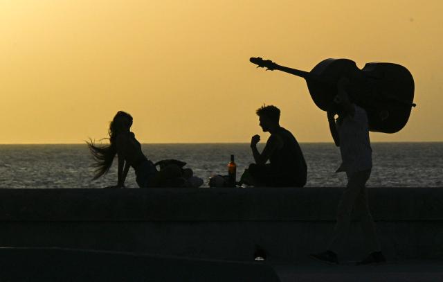 A man carrying a double bass walks past a couple enjoying sunset on the Havana Malecon during a general blackout on July 14, 2026. Cuba's power grid failed for the third time in less than 10 days, sparking another nationwide blackout, the national electricity company said. (Photo by YAMIL LAGE / AFP)