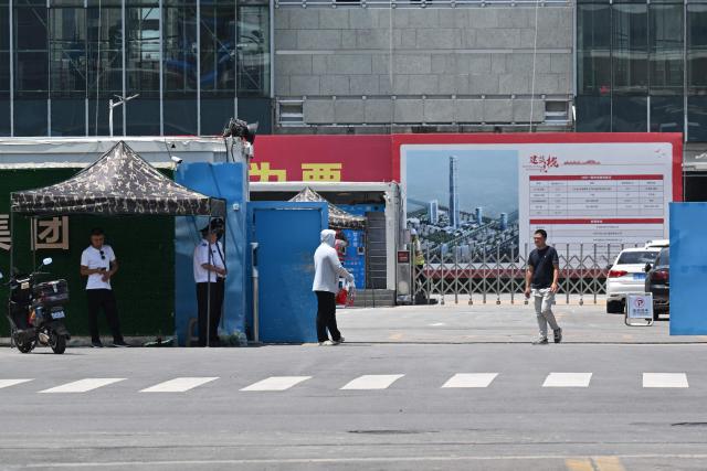 Security personnel (2nd L) keep watch at the entrance to the under-construction Goldin Finance 117 skyscraper in Tianjin on July 14, 2026. The world's tallest unoccupied skyscraper was meant to boast a rooftop swimming pool and a luxury hotel once completed, but instead it has stood deserted in the Chinese port city of Tianjin for years. Work on the soaring Goldin Finance 117 began in 2008, but construction stalled as its developer fell into debt, and the tower remains unfinished 18 years on. (Photo by ADEK BERRY / AFP)