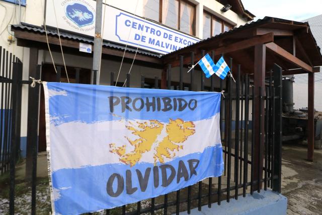 An Argentine national flag featuring a map of the Falkland Islands and a sign reading "Never Forget" is seen at the entrance of the Malvinas War Veterans Center, where fans watch the 2026 World Cup football semi-final match between Argentina and England on a television in Buenos Aires on July 15, 2026. (Photo by Cristian Urrutia / AFP)
