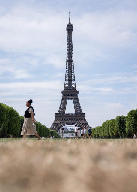 A pedestrian walks past the Eiffel Tower and dried out grass on the Champ de Mars in Paris on July 15, 2026. The Paris region, and large parts of the rest of France, is currently experiencing the third heatwave since May 2026, increasing the risk of fires. The three heatwaves have seen temperature records broken in several countries across Europe and have caused thousands of excess deaths, according to estimates in Belgium, Britain, France and Spain. (Photo by Martin LELIEVRE / AFP)