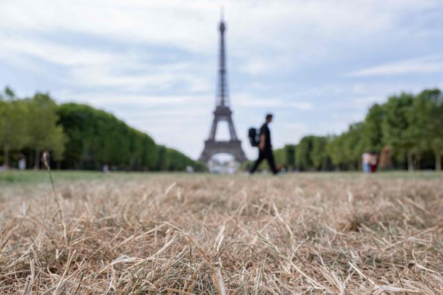 A pedestrian walks past the Eiffel Tower and dried out grass on the Champ de Mars in Paris on July 15, 2026. The Paris region, and large parts of the rest of France, is currently experiencing the third heatwave since May 2026, increasing the risk of fires. The three heatwaves have seen temperature records broken in several countries across Europe and have caused thousands of excess deaths, according to estimates in Belgium, Britain, France and Spain. (Photo by Martin LELIEVRE / AFP)