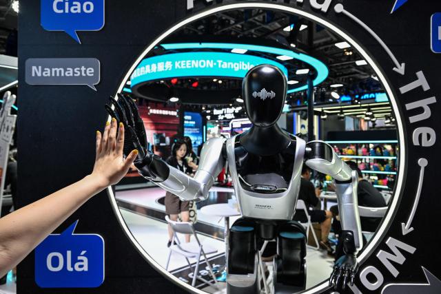 A woman touches the hand of a humanoid robot by Keenon Robotics during the World Artificial Intelligence Conference (WAIC) in Shanghai on July 18, 2026. (Photo by Hector RETAMAL / AFP)