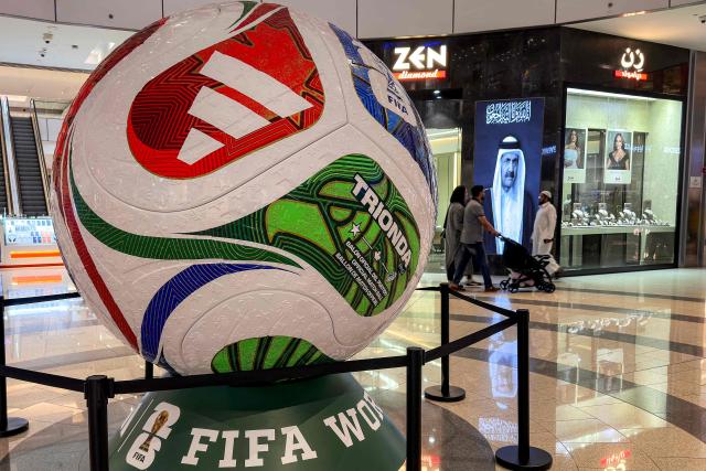 A giant replica of the Trionda, the official match ball for the 2026 FIFA World Cup, is pictured as people walk past a screen displaying an image of Qatar's late former emir Sheikh Hamad bin Khalifa Al-Thani at a shopping mall in Doha on July 18, 2026. (Photo by AFP)