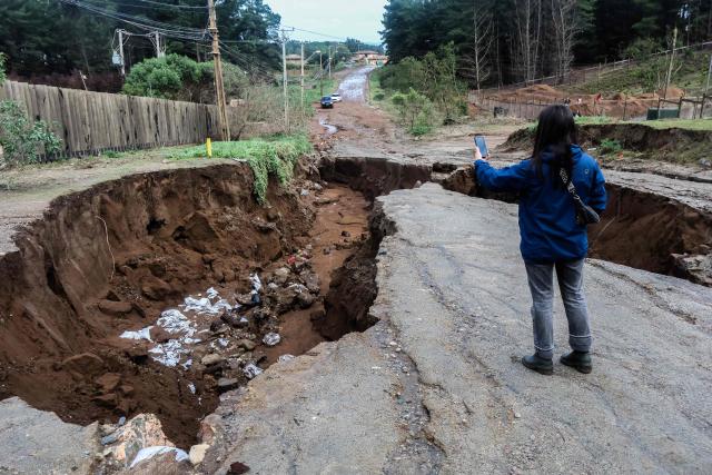 A woman uses a mobile phone as she stands next to a large sinkhole formed after heavy rains in Viсa del Mar, Chile, on July 18, 2026. Three people died and 76 were affected by a storm with heavy rains, swells, and winds that is impacting 10 of the country’s 16 regions, the government reported. (Photo by Cristobal Basaure / AFP)