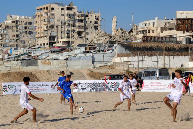 Displaced Palestinians from the Shati Team (blue) and the Gaza Sports Club (White) play in a beach football tournament along the beach in Gaza City on July 18, 2026 with destroyed and damaged buildings pictured in the background. (Photo by Omar AL-QATTAA / AFP)