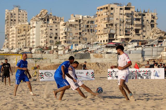 Displaced Palestinians from the Shati Team (blue) and the Gaza Sports Club (White) play in a beach football tournament along the beach in Gaza City on July 18, 2026 with destroyed and damaged buildings pictured in the background. (Photo by Omar AL-QATTAA / AFP)