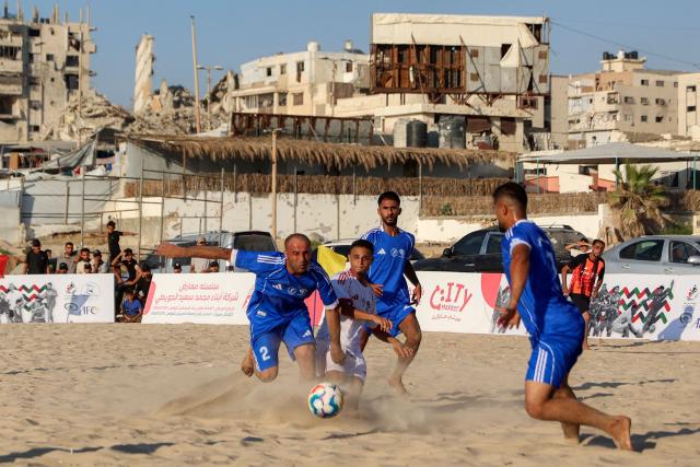 Displaced Palestinians from the Shati Team (blue) and the Gaza Sports Club (White) play in a beach football tournament along the beach in Gaza City on July 18, 2026 with destroyed and damaged buildings pictured in the background. (Photo by Omar AL-QATTAA / AFP)