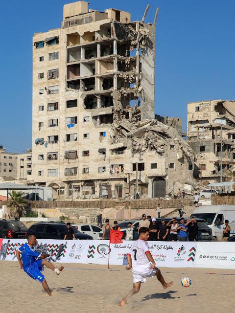 Displaced Palestinians from the Shati Team (blue) and the Gaza Sports Club (White) play in a beach football tournament along the beach in Gaza City on July 18, 2026 with destroyed and damaged buildings pictured in the background. (Photo by Omar AL-QATTAA / AFP)