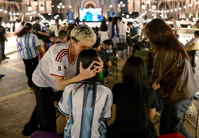 A supporter wearing a jersey of Spain (L) kisses his friend wearing a jersey of Argentina (2nd L) as he celebrates a goal while watching a live broadcast of the 2026 World Cup football final match between Spain and Argentina on a screen at a park in Phnom Penh on July 20, 2026. (Photo by TANG CHHIN Sothy / AFP)