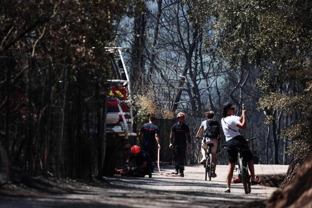 Firefighters stand next to their vehicle as a cyclist takes images with smartphone following a wildfire in Taradeau, in Var department of Provence-Alpes-Côte d'Azur region, southeastern France on July 20, 2026. The intense fire that broke out on July 19 afternoon in the Var, was showing signs of improvement by evening, but had affected around ten houses and around fifty houses were affected. (Photo by Thibaud MORITZ / AFP)