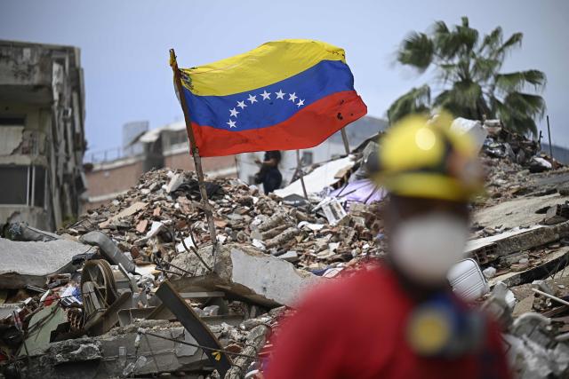 A Venezuelan flag flies over the rubble of collapsed buildings as a rescue worker stands in the foreground in Caraballeda, Venezuela, on July 20, 2026, following the earthquakes that struck the area. (Photo by Federico PARRA / AFP)