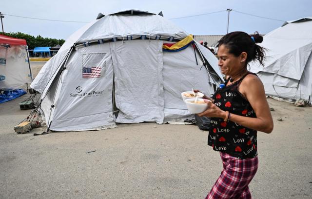 A woman carries food past a tent with a US flag in Catia La Mar, Venezuela, on July 20, 2026. The number of people killed in the powerful dual Venezuela earthquakes last month rose to 5,278, according to official figures updated Monday. The June 24 earthquakes, which recorded magnitudes of 7.2 and 7.5, profoundly impacted the north of the country, particularly the coastal state of La Guaira, near the capital Caracas. (Photo by Juan BARRETO / AFP)
