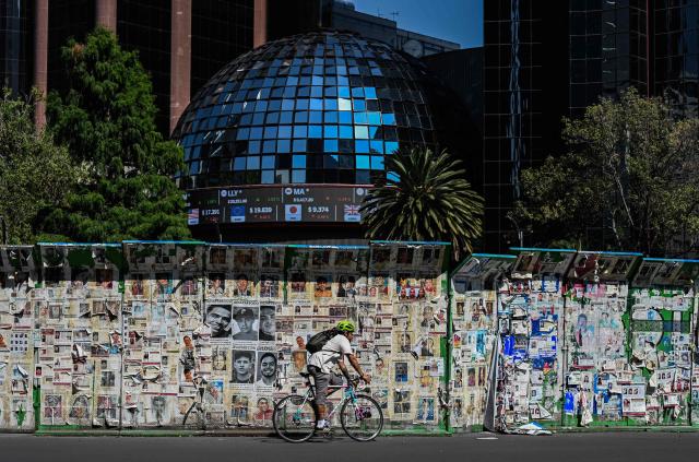 A man rides a bicycle past a mural covered with photographs of missing persons while a screen on the Stock Exchange building displays currency exchange rates in Mexico City on July 21, 2026. (Photo by Yuri CORTEZ / AFP)