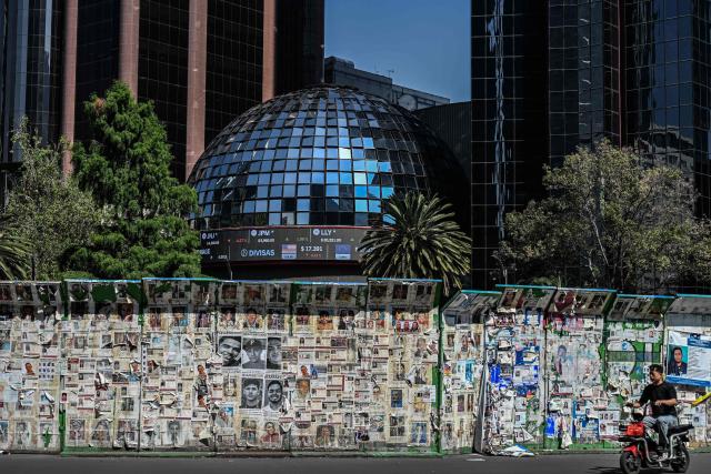 A man rides a motorcycle past a mural covered with photographs of missing persons while a screen on the Stock Exchange building displays currency exchange rates in Mexico City on July 21, 2026. (Photo by Yuri CORTEZ / AFP)