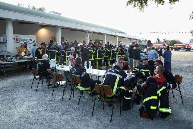 Firefighters and inhabitants take their breakfast together outside the Gerard Blanc festival hall in Le Porge, southwestern France on July 23, 2026, following a wildfire and the evacuation of the residents. A highly virulent wildfire has swept through more than 2,000 hectares since July 22, 2026 near the Arcachon Basin (Gironde), where nearly 12,000 residents and tourists have been evacuated in total from this area of the Landes de Gascogne forest, already devastated by flames in 2022. (Photo by ROMAIN PERROCHEAU / AFP)