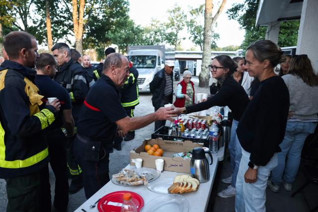 Firefighters and inhabitants take their breakfast together outside the Gerard Blanc festival hall in Le Porge, southwestern France on July 23, 2026, following a wildfire and the evacuation of the residents. A highly virulent wildfire has swept through more than 2,000 hectares since July 22, 2026 near the Arcachon Basin (Gironde), where nearly 12,000 residents and tourists have been evacuated in total from this area of the Landes de Gascogne forest, already devastated by flames in 2022. (Photo by ROMAIN PERROCHEAU / AFP)