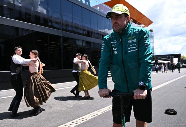 Aston Martin's Spanish driver Fernando Alonso (R) passes by dancers in traditional Hungarian costumes prior to the first practice session at the Hungaroring racetrack in Mogyorod near Budapest, Hungary, on July 24, 2026 ahead of the Formula One Hungarian Grand Prix. (Photo by Attila KISBENEDEK / AFP)
