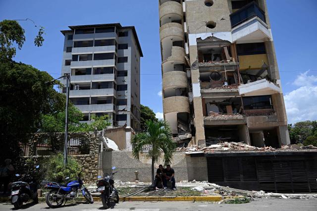 Two people sit in the shade of a tree in front of an apartment building damaged by twin earthquakes in Catia La Mar, La Guaira state, Venezuela, on July 24, 2026, one month after the tremors struck the area. Recovering from the earthquakes in Venezuela will take years, the Red Cross said on July 24, one month on from the June 24 disaster that killed more than 5,000 people. (Photo by Federico PARRA / AFP)