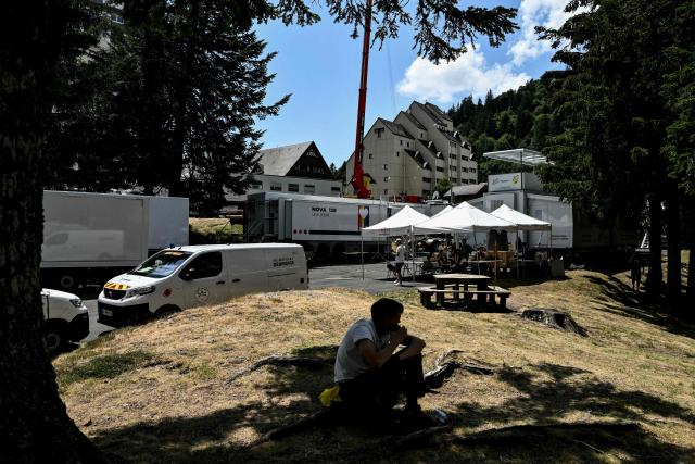 A worker seeks shade by logistical trucks parked tightly together in the "Zone Technique" of the finish line area of the 10th stage of the 113th edition of the Tour de France cycling race, 166,6 km between Aurillac and Le Lioran in the Massif Central, on July 14, 2026. Hours before the Tour de France peloton arrives, an empty field or car park on the edge of town is already being transformed.
Beyond the crowds, barriers and television cameras, a temporary settlement rises each day in the race's finish town. In a field, industrial estate or patch of open ground adjacent to the stages finish line, designated as the Zone Technique (Technical Zone), hundreds of workers transform an empty space into a functioning village capable of supporting one of the world's largest sporting events. Then, less than 24 hours later, it is dismantled and moved on.
Much of the operation unfolds overnight. In the early hours, trucks stream into the Zone Technique from the previous finish town, parking with military precision as crews unload equipment and connect power supplies before dawn. Many workers live on a shifted timetable, sleeping when they can during the afternoon in camping chairs, hamacs or shaded corners of the compound before another night of logistics begins.
For three weeks each July, the Tour carries far more than cyclists across France. Technicians, builders, media crews, podium hosts and hostesses travel with the race, bringing their own power, communications, food services and sanitation infrastructure.
Together, they form a population that descends daily on towns and villages of every size, bringing with them their own infrastructure, services and routines.
Seen from inside the Zone Technique, the Tour de France is not only a cycling event. It is a nomadic settlement crossing the country, rebuilding itself from scratch every day, wherever the finish line happens to be. (Photo by Jeff PACHOUD / AFP)