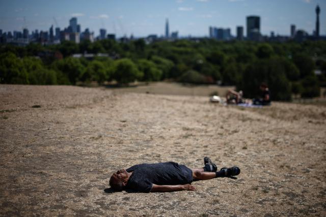 A man lies down on the dry, sun-scorched grass of Primrose Hill, backdropped by the skyline of central London, on July 27, 2026. Temperatures were forecast this to rise back into the 30s for parts of England, following three heatwaves so far over the summer. (Photo by Henry Nicholls / AFP)