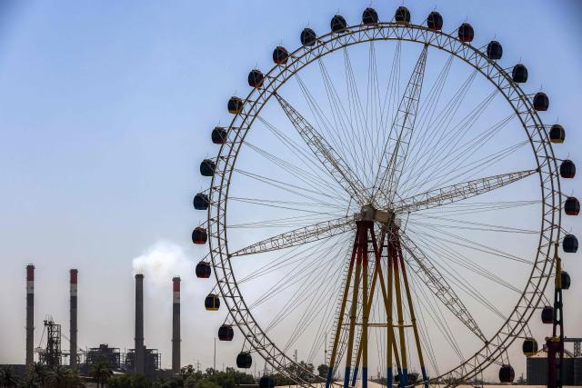 A large Ferris wheel is pictured near the Dora (Daura) Thermal Power Station in the Dora district of southern Baghdad on July 27, 2026. In oil-rich Iraq, many households have just a few hours of state electricity per day, and those who can afford it use private generators to keep fridges and air conditioners running. The country is the second-largest oil producer in the OPEC cartel, but despite its immense oil and gas reserves, it remains dependent on imports to meet its electricity needs. (Photo by AHMAD AL-RUBAYE / AFP)
