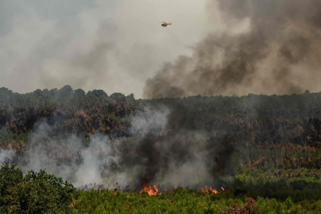 A helicopter flies over smoke rising from a forest fire outside Lege-Cap-Ferret, southwestern France on July 28, 2026. Over the past six days, the wildfire near Bordeaux has ravaged 42,000 hectares (103,000 acres), forced 220,000 people to flee and destroyed 240 houses. Firefighters said on July 28, 2026 the fire had not expanded overnight as they managed to tackle several flareups. (Photo by Ed JONES / AFP)
