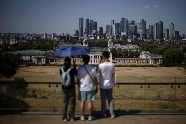 People shelter from the sun as they look out at the Old Royal Naval College (behind C) and the skyline of Canary Wharf, across the dried out and yellow grass in Greenwich Park, south-east London on July 29, 2026. Just over half of England has been declared in drought after "record-low rain and exceptionally high temperatures", the UK government said on July 29. (Photo by Henry NICHOLLS / AFP)