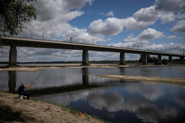 A fisherman sits by the river Danube, in front of the Calafat-Vidin bridge, connecting Bulgaria and Romania near the village Antimovo, southeastern Bulgaria, on July 22, 2026. At the Romanian town of Corabia on the banks of the Danube river, some boats are stranded in shallow water full of algae in the touristic port, which lies deserted as ships can no longer dock due to sandbars (Photo by Nikolay DOYCHINOV / AFP)