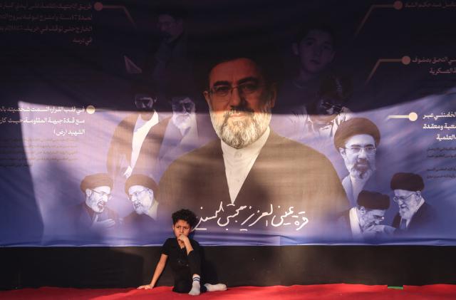 A boy sits beneath a banner depicting Iranian supreme leader Mojtaba Khamenei as Shia pilgrims march through Baghdad's Dora district towards Karbala to take part in the Arbaeen pilgrimage on July 30, 2026. The Arbaeen marks one of the most significant events in the Shia Muslim calendar, during which pilgrims, many from Iran, flock to Iraq to visit the holy shrines. (Photo by AHMAD AL-RUBAYE / AFP)