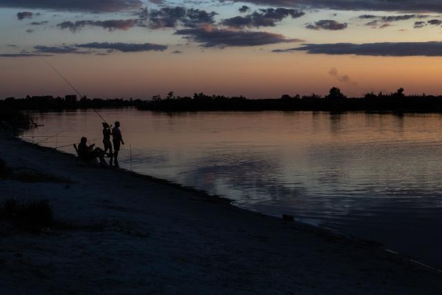 People fish in a river outside the city of Dnipro as smoke rises in the distance during a Russian air attack on July 30, 2026, amid the Russian invasion of Ukraine. (Photo by Roman PILIPEY / AFP)