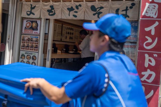 A man eats noodles in a shop as a delivery employee from Sagawa, wearing a specialised jacket with a fan to cool down, walks down a road to deliver packages in Tokyo on July 30, 2026. (Photo by Andrew CABALLERO-REYNOLDS / AFP)