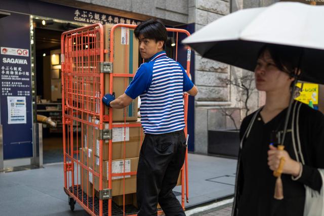 A delivery employee from Sagawa moves a crate of boxes for delivery as a woman uses an umbrella for shade from the sun in Tokyo on July 30, 2026. (Photo by Andrew CABALLERO-REYNOLDS / AFP)