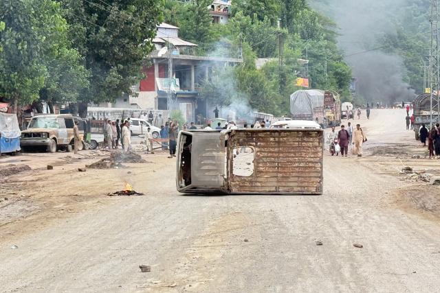 People walk along a street after protestors blocked a road with the wreckage of a vehicle following clashes with police in Muzaffarabad on July 31, 2026 amid unrest during the first phase of voting for the local legislature in Pakistan-administered Kashmir. Protesters and security personnel engaged in deadly clashes in Pakistan-administered Kashmir as the sensitive Himalayan region goes to the polls, an official said on July 28. (Photo by Sajjad QAYYUM / AFP)