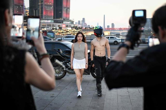 This picture taken on July 20, 2026 shows a woman posing with a motorcycle rider offering photo and video services along a street in Chongqing, southwestern China. The roar of engines fills the night air in China's Chongqing city as tourists awkwardly clamber onto gleaming motorbikes and cling to muscular drivers to star in personalised videos. (Photo by WANG Zhao / AFP) / To go with 'CHINA-MOTORBIKE-INTERNET,FOCUS' by Sam DAVIES