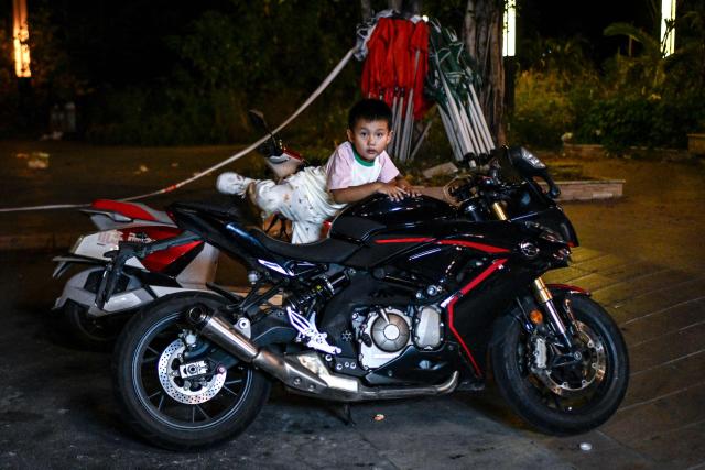 This picture taken on July 21, 2026 shows a child climbing onto a motorcycle at a riding photo and video service site in Chongqing, southwestern China. The roar of engines fills the night air in China's Chongqing city as tourists awkwardly clamber onto gleaming motorbikes and cling to muscular drivers to star in personalised videos. (Photo by WANG Zhao / AFP) / To go with 'CHINA-MOTORBIKE-INTERNET,FOCUS' by Sam DAVIES