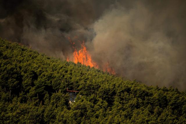 TOPSHOT - This photograph shows wildfire flames and heavy smoke rising above a house from a wildfire in the area of Porto Germeno, some 70 kilometres (45 miles) northwest of Athens in the region of Viotia, on August 2, 2026. Greece on August 2, 2026, faced multiple fronts and a growing damage toll after a week of wildfires devastated over 12,000 hectares of forest and agricultural land, experts said. Porto Germeno and other villages were evacuated when the fire broke out on July 31, but local authorities say scores of homes were damaged or destroyed. Hit hard by the climate crisis like the rest of the Mediterranean region, the country is plagued by wildfires every summer due to high temperatures, frequent heatwaves, and drought. (Photo by Angelos TZORTZINIS / AFP)