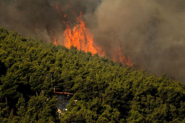 This photograph shows wildfire flames and heavy smoke rising above a house from a wildfire in the area of Porto Germeno, some 70 kilometres (45 miles) northwest of Athens in the region of Viotia, on August 2, 2026. Hundreds of firefighters in Greece battle to save a popular coastal resort northwest of Athens as violent winds continued to lash the country, hampering operations. Hit hard by the climate crisis like the rest of the Mediterranean region, the country is plagued by wildfires every summer due to high temperatures, frequent heatwaves, and drought. (Photo by Angelos TZORTZINIS / AFP)