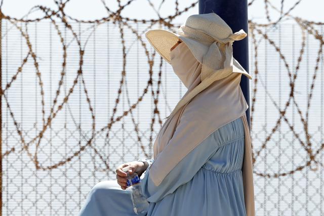 A woman sits by a barbed-wire fence as relatives and friends wait for the return of people who had previously crossed into Spain's North African enclave of Ceuta, at Morocca's Bab Sebta border crossing on the outskirts of Fnideq on August 2, 2026. As many as 60,000 migrants entered Ceuta in recent days, mostly swimming around the small border post that juts into the Mediterranean Sea, in an unprecedented surge that prompted an international crisis for Spain. The mass crossing into the tiny Spanish territory resulted in at least 67 deaths, according to Spanish authorities, and led to clashes with Moroccan police on the shared border. (Photo by Abdel Majid BZIOUAT / AFP)